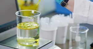 A glass container with yellow liquid being dropped, a scientific lab can be seen behind.